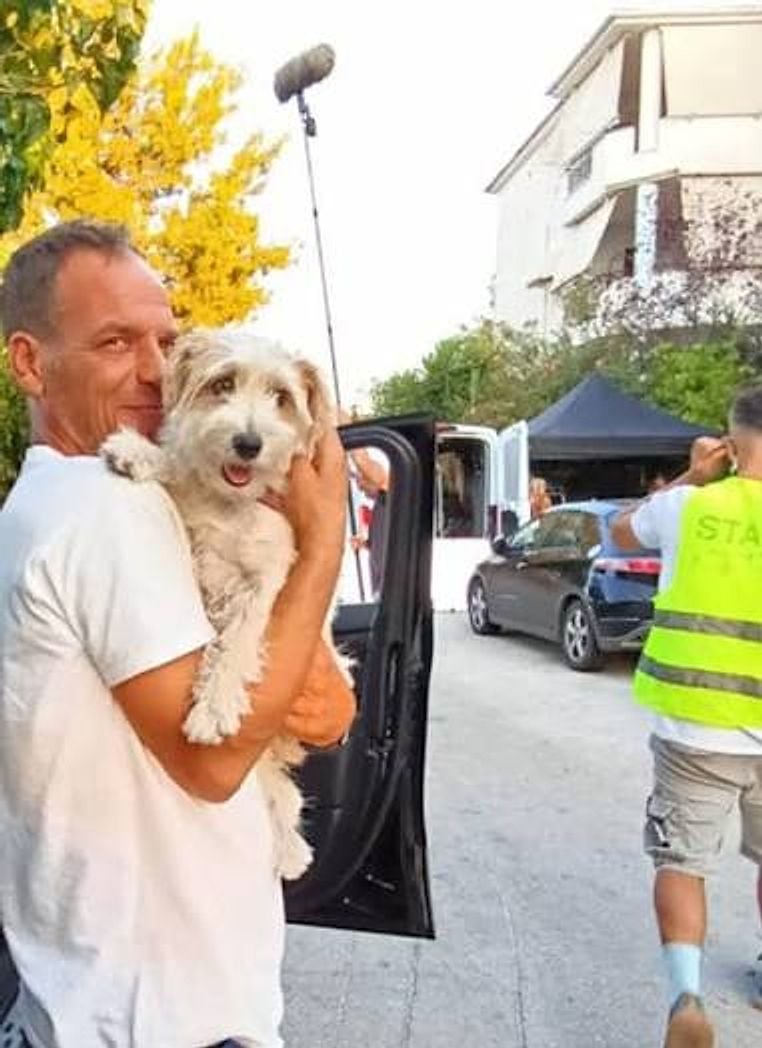 Στα παρασκήνια γυρίσματος: εκπαιδευτής κρατά αγκαλιά ένα λευκό σκυλάκι, με το μικρόφωνο από πάνω